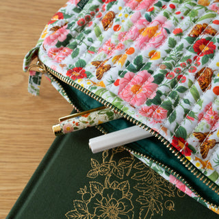 A close-up of a quilted fabric zip pouch with a multicolor floral and butterfly print, shown partially open on a wooden surface with pens and a green notebook peeking out.