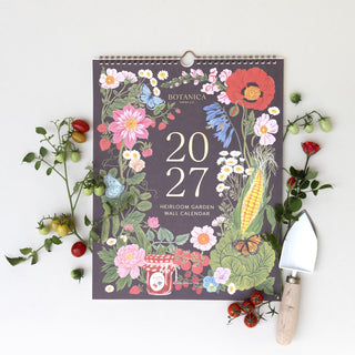 A 2027 Herloom Garden Wall Calendar by Botanica is displayed in a flat-lay arrangement on a white background, surrounded by fresh garden elements including cherry tomatoes, a small gardening trowel, and a hanging wire loop at the top of the calendar.