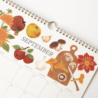 A spiral-bound September wall calendar page with autumn-themed illustrations including apples, mushrooms, garlic, flowers, and a cutting board, photographed on a white background.