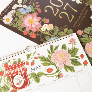 A close-up of a spiral-bound heirloom garden wall calendar showing the May page with floral illustrations, strawberries, a pink flower, and a red-and-white checkered jar graphic. Another view in the background shows a dark brown cover with gold '2027' text and more botanical artwork.