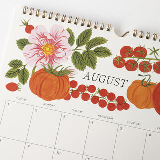 A close-up of a spiral-bound August wall calendar page with a white background and autumn harvest illustration, including tomatoes, cherry tomatoes, and a pink rose.