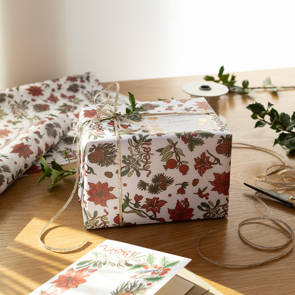 Gift box wrapped in Christmas floral paper on a wooden table with decorative items. Art by Oana Befort for Botanica Paper Co.