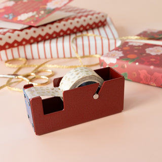 A small dark red tape dispenser holding a roll of patterned washi tape, styled with gift boxes, wrapping paper, and gold ribbon on a soft peach background.