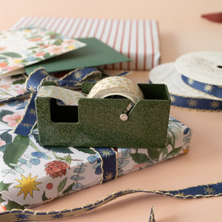 A dark green textured tape dispenser holding a roll of cream tape with small gold stars, styled on top of floral wrapping paper with ribbed gift boxes and ribbon spools in the background.