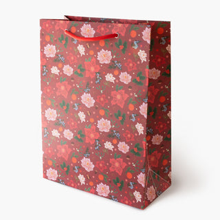 A red gift bag with a festive floral pattern featuring pink poinsettias, green leaves, and orange berries, complete with red paper handles, set against a plain white background.