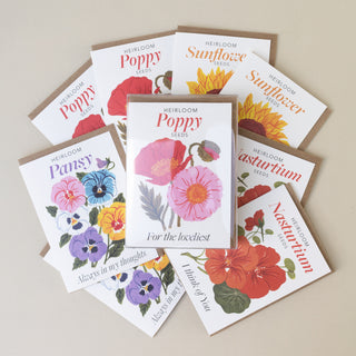 A fan arrangement of illustrated seed packet cards for heirloom seeds, including poppy, sunflower, pansy, nasturtium, and marigold, with brown envelopes behind them on a light beige background.