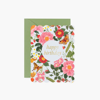 A floral birthday card with a scalloped edge, featuring pink flowers, green leaves, butterflies, and the words 'happy birthday' in gold script, shown with a sage green envelope behind it on a light gray background.