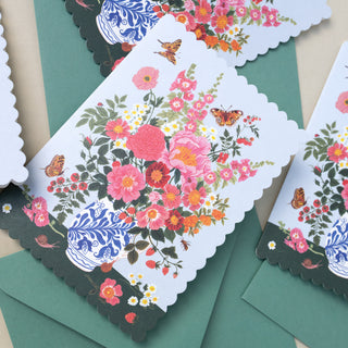 A close-up of a decorative paper card with scalloped edges, featuring a colorful floral bouquet, butterflies, and a blue-and-white porcelain vase. The card is layered over green envelopes on a neutral surface.