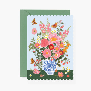 A floral greeting card with a scalloped white border, featuring a vibrant bouquet of pink, red, and orange flowers in a blue-and-white patterned vase, shown with a sage green envelope on a light gray background.