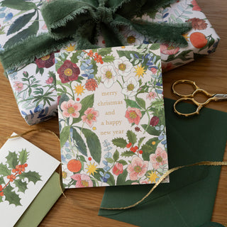 A Christmas greeting card with a botanical floral border and gold text reading 'merry christmas and a happy new year' is styled on a wooden surface with wrapping paper, a green ribbon, gold scissors, and a small holly tag.
