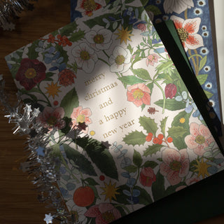 A floral Christmas greeting card with the text 'merry christmas and a happy new year' in gold, styled on a wooden surface with silver tinsel and a dark green book or notebook underneath.