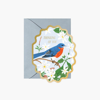 A decorative greeting card with a scalloped gold border, featuring a bluebird perched among white flowers and green leaves, with the words 'THINKING OF YOU' in gold. The card is shown with a light blue envelope behind it on a plain white background.