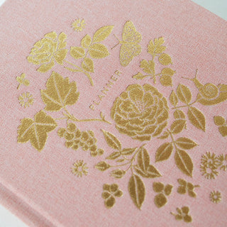 Macro detail of Reverie planner cover showing the luxurious gold foil lush floral stamping technique and premium soft pink book linen cloth texture, demonstrating Botanica Paper Co.'s craftsmanship