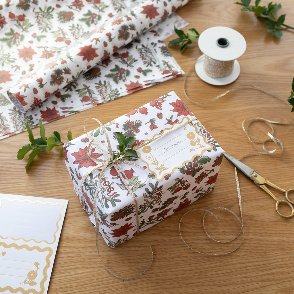 Gift wrapped in Christmas floral paper with a label, surrounded by gift wrap, scissors, and ribbons on a wooden surface.