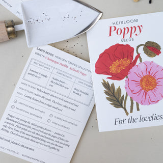 A printed seed packet for Heirloom Poppy Seeds lies on a light beige surface next to a small metal trowel. The packet features large illustrations of red and pink poppy flowers with green leaves, and the front text reads 'Heirloom Poppy Seeds For the lovelies.