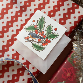 A square white Christmas greeting card featuring an illustration of a bluebird on a holly branch with red berries, placed on top of a translucent white envelope over red patterned gift wrapping paper.