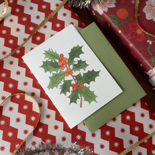 A white Christmas greeting card featuring a watercolor illustration of holly leaves and red berries, placed on top of a green envelope. The card is styled on a red and white patterned holiday background with gift wrapping and gold ribbon.