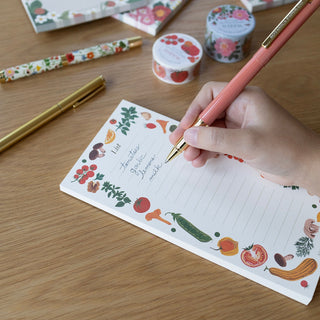 A hand writing in a lined notepad with a vegetable-themed border, using a pink pen with gold trim on a wooden desk. Other floral stationery items are arranged nearby.