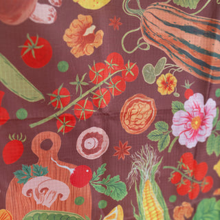 A close-up of a printed ripstop shopping bag featuring a colorful vegetable and fruit pattern on a dark plum-brown background, including tomatoes, cucumbers, corn, mushrooms, and flowers.