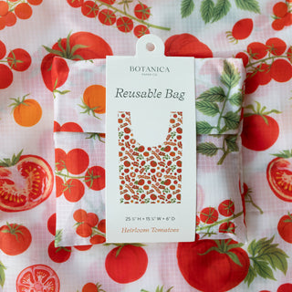 A Botanica reusable bag in a hotloom tomato print, shown folded with its white cardboard retail tag attached. The background is a matching flat-lay of the same tomato-patterned fabric.