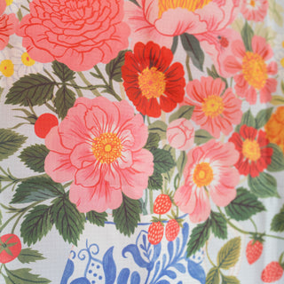 A close-up of a floral fabric print featuring large pink and red flowers, green leaves, and small red berries on a white background with a blue pattern at the bottom.
