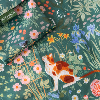 A close-up of a botanical reusable bag featuring a ginger cat, pink flowers, blue irises, and green leaves on a dark teal-green background. A folded matching pouch with a 'BOTANICA FRESH CO.