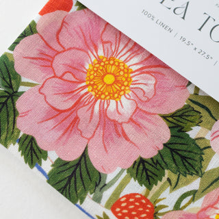 A close-up of a floral printed linen fabric with a large pink flower, green leaves, and a strawberry, with part of a white paper label visible reading '100% LINEN' and '19.5" x 27.
