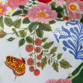 A close-up of a white linen towel fabric with a colorful botanical print featuring pink flowers, green leaves, red tomatoes, a yellow-and-brown butterfly, and a blue ornamental pattern.