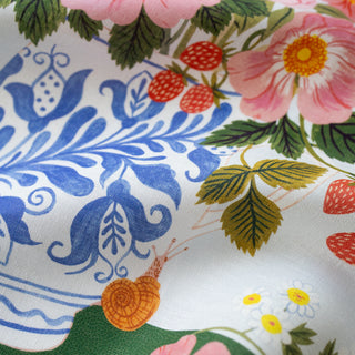 A close-up of a linen tea towel  fabric with a colorful botanical print featuring blue floral motifs, pink flowers, green leaves, red strawberries, and a small orange snail.