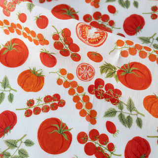 A close-up of linen tea towel fabric with a vibrant tomato print, featuring whole red tomatoes, sliced tomatoes, clusters of cherry tomatoes, and green leaves.