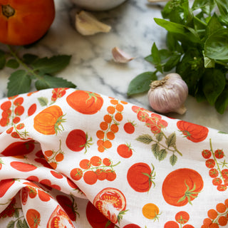 A white linen kitchen towel  printed with a colorful tomato and cherry tomato pattern, styled on a marble surface with fresh basil, garlic, and a whole tomato in the background.