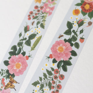 Two narrow decorative paper tape strips with a botanical print, laid diagonally on a white textured surface. The pattern features pink flowers, green leaves, red berries, small yellow daisies, and a butterfly.
